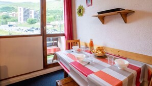 A dining area with a table set for two, a window with a view of buildings and greenery, and a wall-mounted shelf above the table.