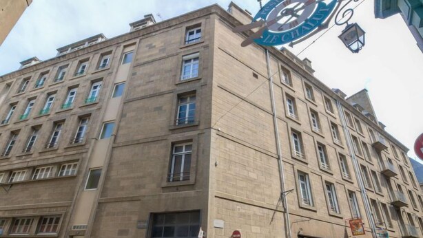 Un edificio de varios pisos con un reloj llamativo en la fachada, una puerta principal cerrada y un pequeño toldo sobre la entrada.