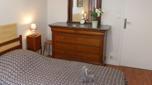 Un dormitorio con un aparador de madera, una cama con colcha estampada, una mesita de noche con lámpara, una pequeña mesa con un reloj y una puerta con picaporte.