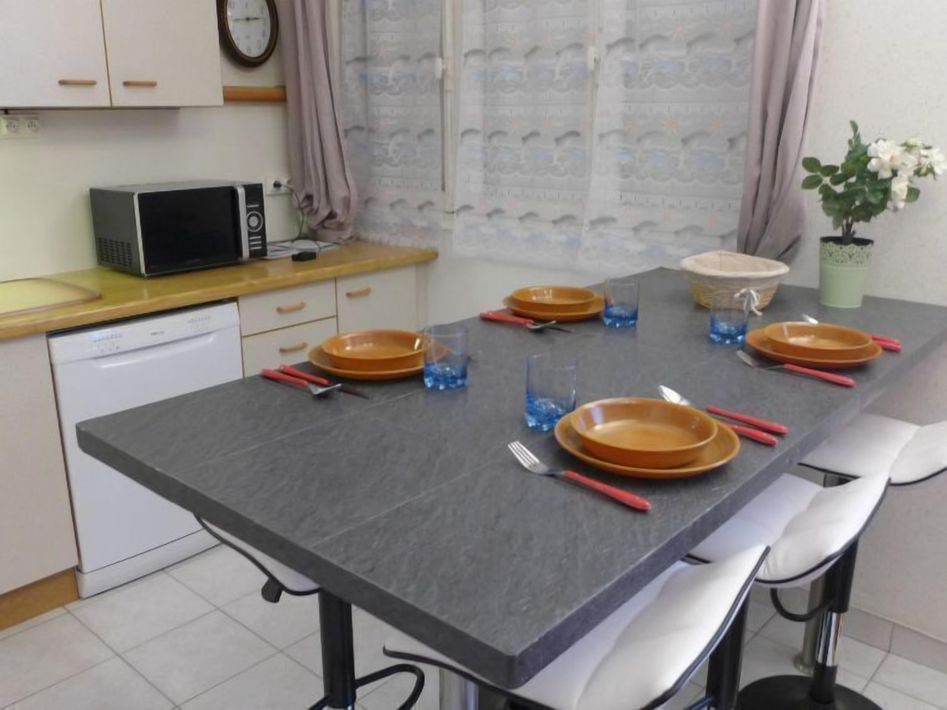Une cuisine avec un plan de travail gris, des placards blancs, un four à micro-ondes et un coin repas avec une table dressée pour quatre personnes.