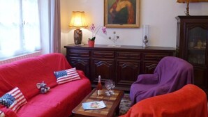 Una sala de estar con un sofá rojo, un sillón morado, una mesa de centro de madera con una botella y vasos, y un cuadro en la pared.
