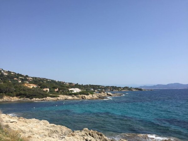 A coastal landscape with rocky shores, clear blue sea, and a hillside with buildings.