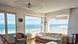 Een ruime woonkamer met grote ramen met uitzicht op een strand, houten vloeren, een groene fauteuil, een beige bank en een houten dressoir.