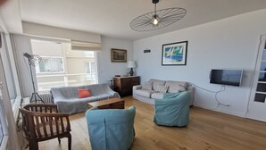 Een woonkamer met een bank, twee blauwe fauteuils, een houten stoel, een salontafel, een vloerlamp, een televisie en kunstwerken aan de wanden.