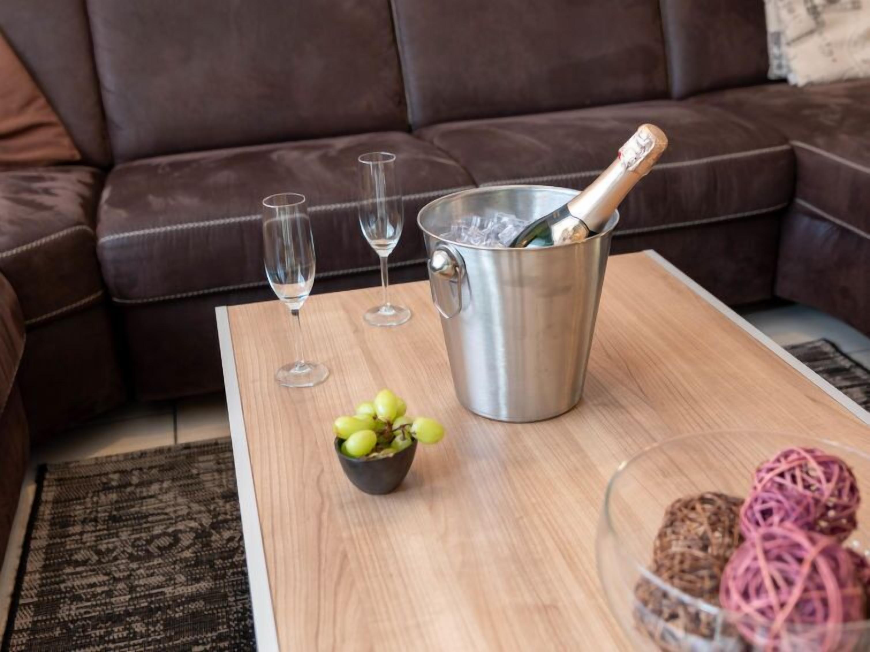 Ein gemütliches Wohnzimmer mit einem braunen Ledersofa, einem Holztisch, einem Metall-Eimer mit einer Champagnerflasche, zwei Weingläsern und einer Schüssel mit Trauben.