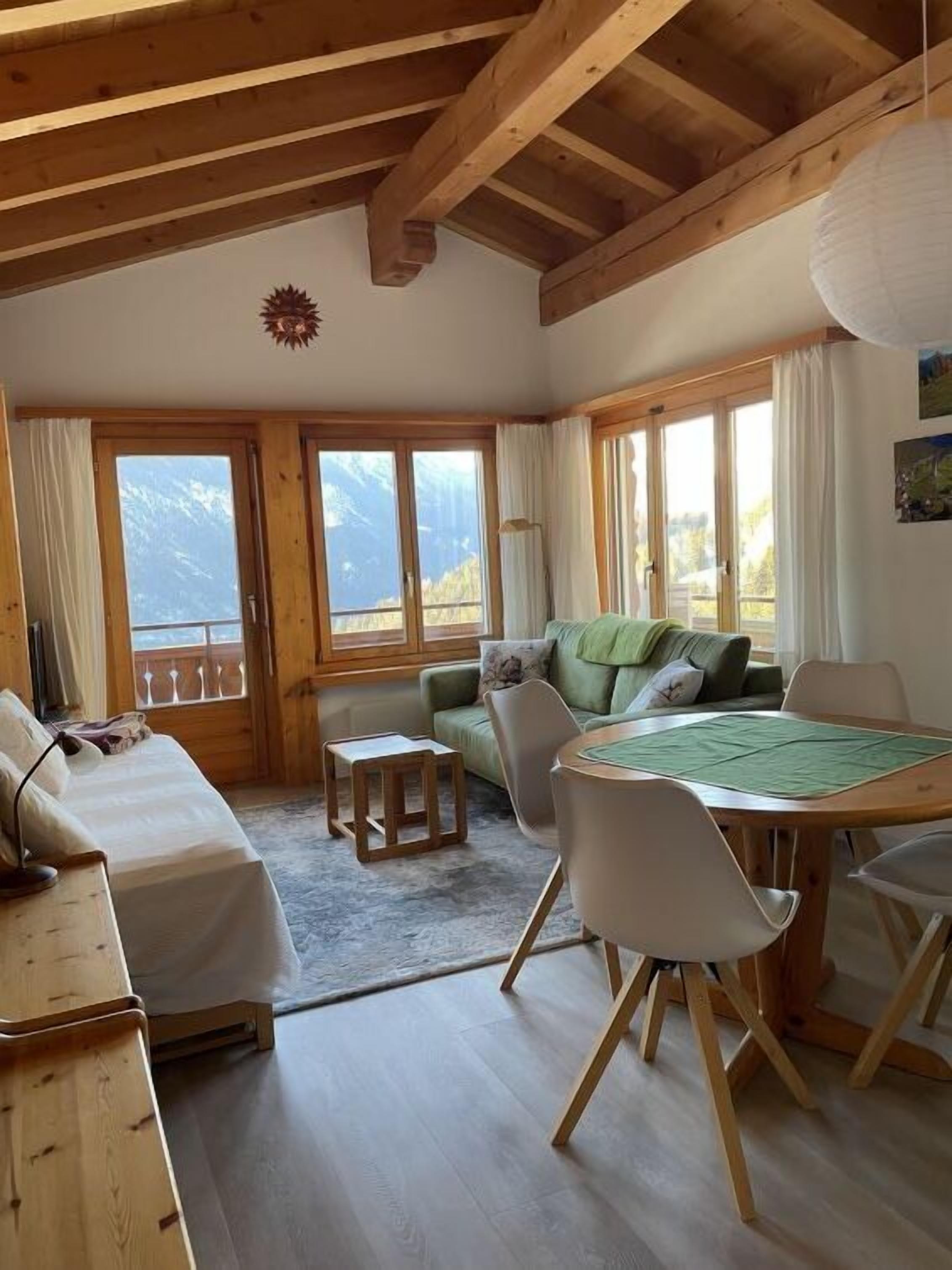 Une pièce chaleureuse avec un plafond en bois, un canapé, une table à manger avec des chaises et de grandes fenêtres offrant une vue sur les montagnes.