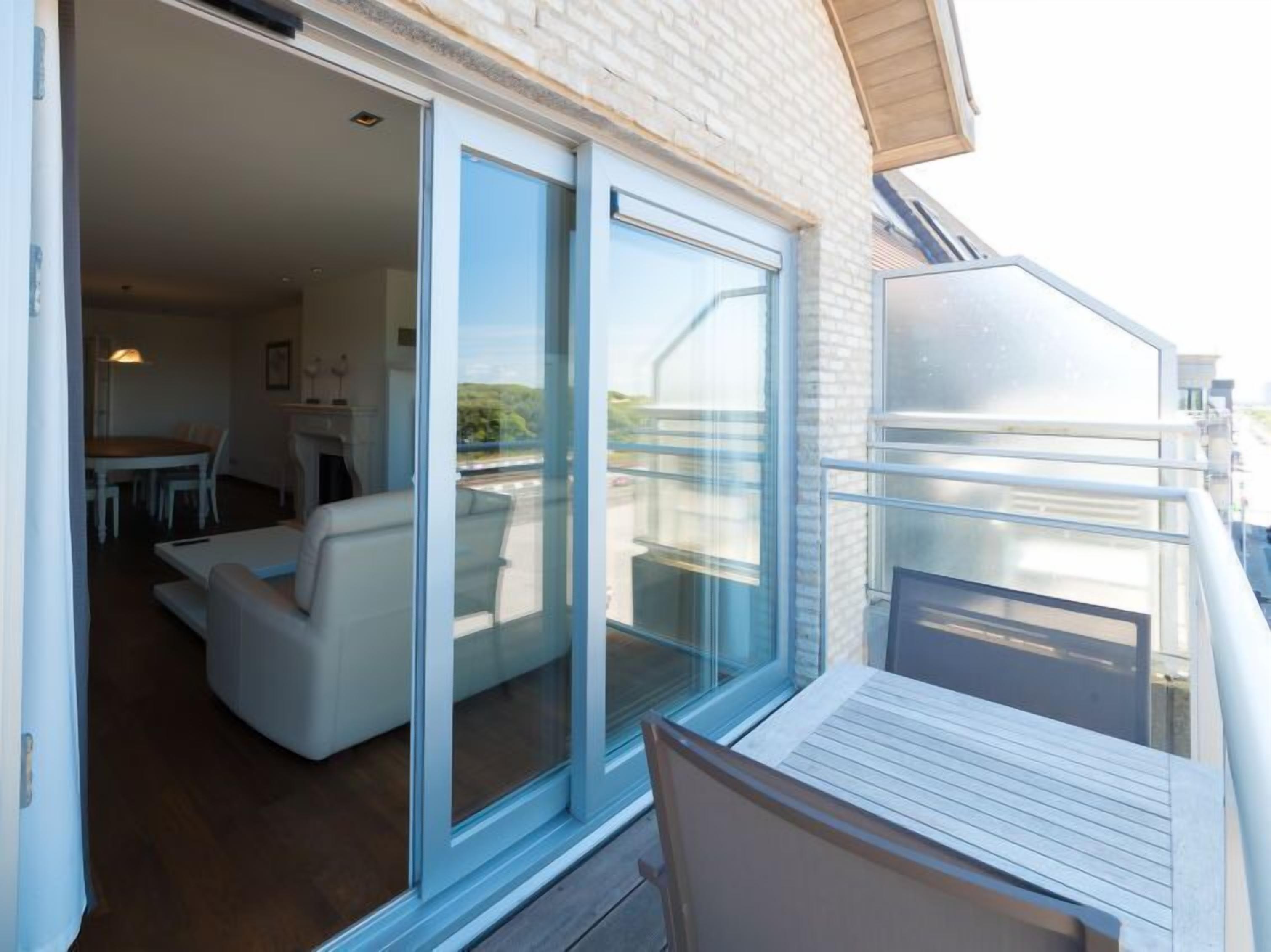 Un balcon avec une table et des chaises en bois, une porte-fenêtre coulissante donnant sur un espace de vie comprenant un canapé et une table à manger.
