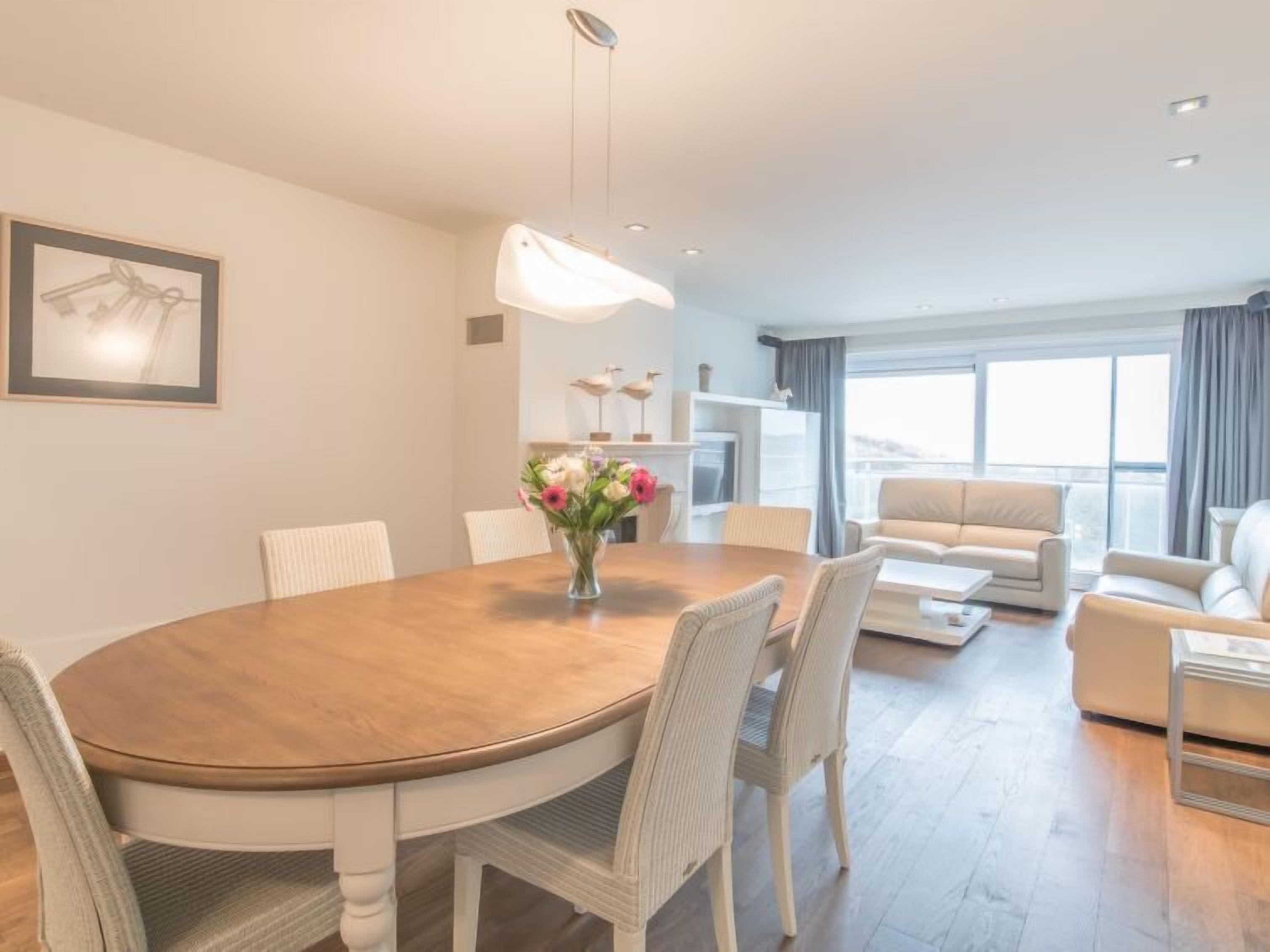 Una sala de estar moderna con una mesa de comedor, sillas de mimbre blanco, un sofá y un ventanal con cortinas.