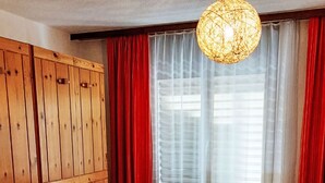Huoneessa on puinen vaatekaappi, suuri ikkuna, jossa on ohuet verhot, sekä katosta roikkuva valaisin.