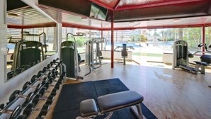 Academia com diversos equipamentos de exercício, um banco e vista para uma piscina e palmeiras.