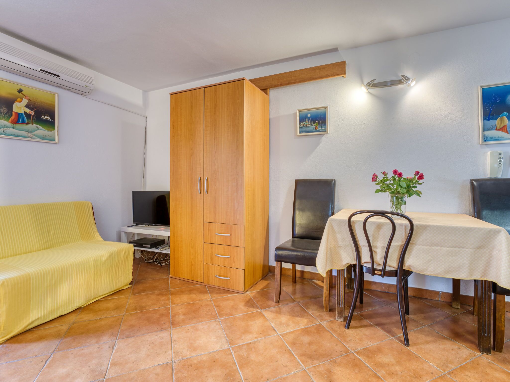 A room with a yellow sofa, a dining table with chairs, a wooden wardrobe, a television, and a painting on the wall.
