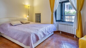 Un dormitorio con cama, mesita de noche, lámpara, ventana con cortinas y radiador.