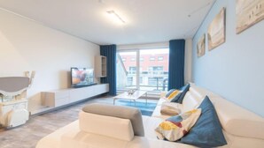 En moderne stue med en hvid sofa, et fladskærms-tv og et stort vindue med udsigt over byen.