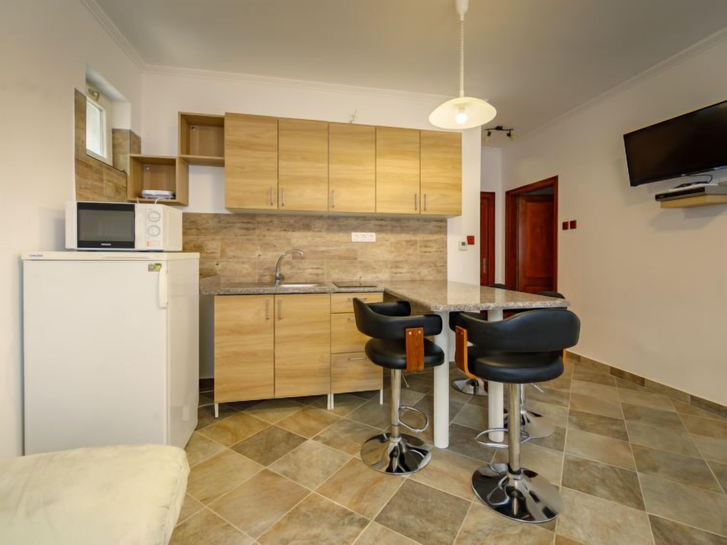 Cocina pequeña con armarios de madera, microondas, lavamanos y dos taburetes de bar.