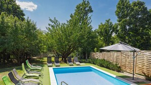 Una piscina rectangular con una escalera metálica, rodeada de césped artificial verde. Se observan varias sillas de descanso y una valla de madera al fondo.