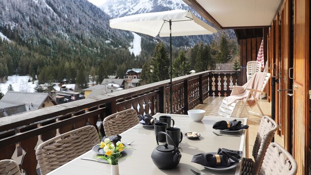 Un comedor con una mesa preparada para dos personas, un florero con flores, una tetera negra y vistas de montañas nevadas.