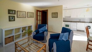 Una sala de estar con dos sofás, una mesa de centro y un mueble para televisión. Se ve una cocina con gabinetes y electrodomésticos blancos al fondo.