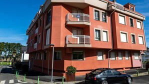Un edificio residencial de varios pisos con balcones, un coche estacionado en frente y un cielo azul despejado.