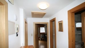 A narrow hallway with wooden flooring, white walls, and a ceiling light. There is a wooden bench on the left, a door on the right, and a small table with a telephone on the wall.