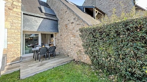 A stone building with a sloped roof, a wooden deck, and a glass door leading to an interior space with a table and chairs. - Les Alizés by Interhome (Carnac)