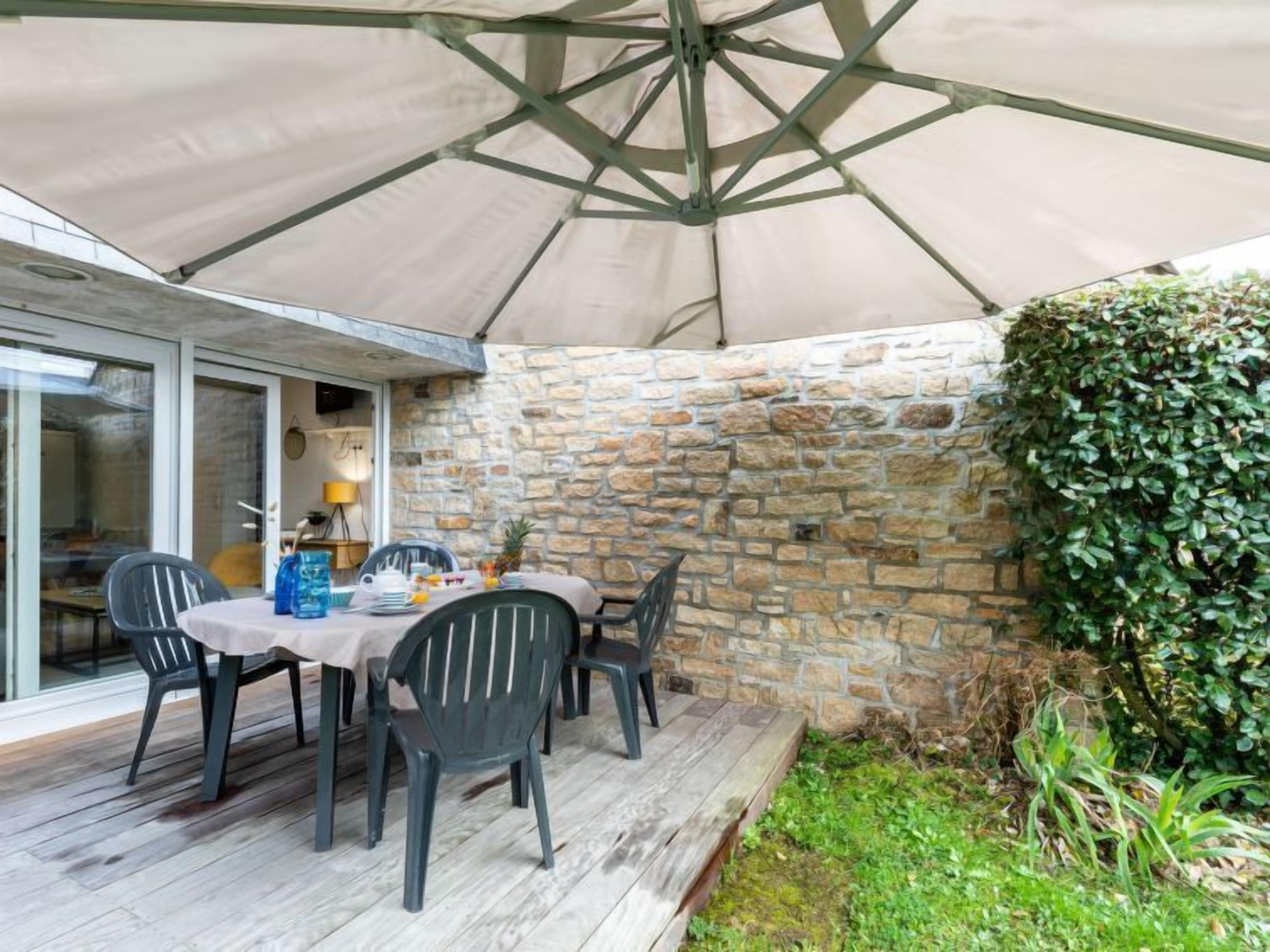 A covered patio with a table set for four, surrounded by stone walls and a green hedge.