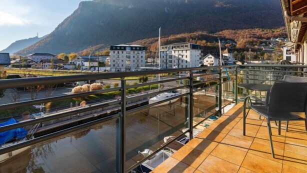 A balcony with a view of a river, boats, and buildings in the background. - La Perouse D1/2 by Interhome (Le Bouveret)