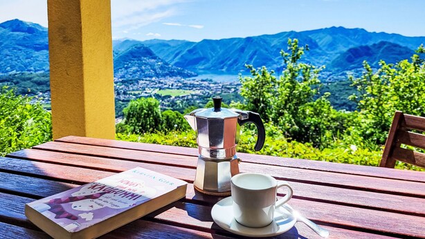 Ein Tisch mit einem Buch, einer Kaffeekanne und einer Tasse auf einer hölzernen Bank, im Hintergrund ein schöner Bergblick.