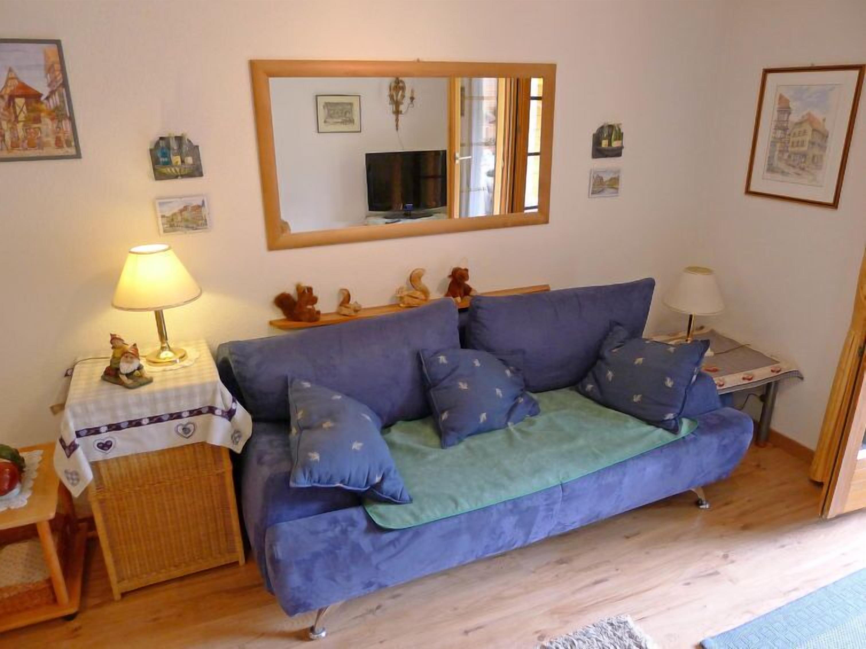 Een woonkamer met een blauwe bank, een bijzettafeltje met een lamp, een rieten bijzettafeltje en een spiegel aan de muur.