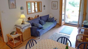 Een gezellige woonkamer met een blauwe bank, een klein tafeltje met een lamp, en een eethoek met stoelen en een tafel.