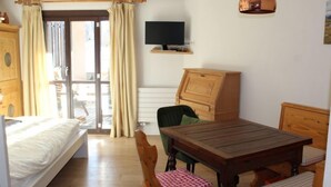 Ein gemütliches Zimmer mit einem Bett, einem Holztisch, einem Stuhl, einem Fernseher und einem Fenster mit Vorhängen.