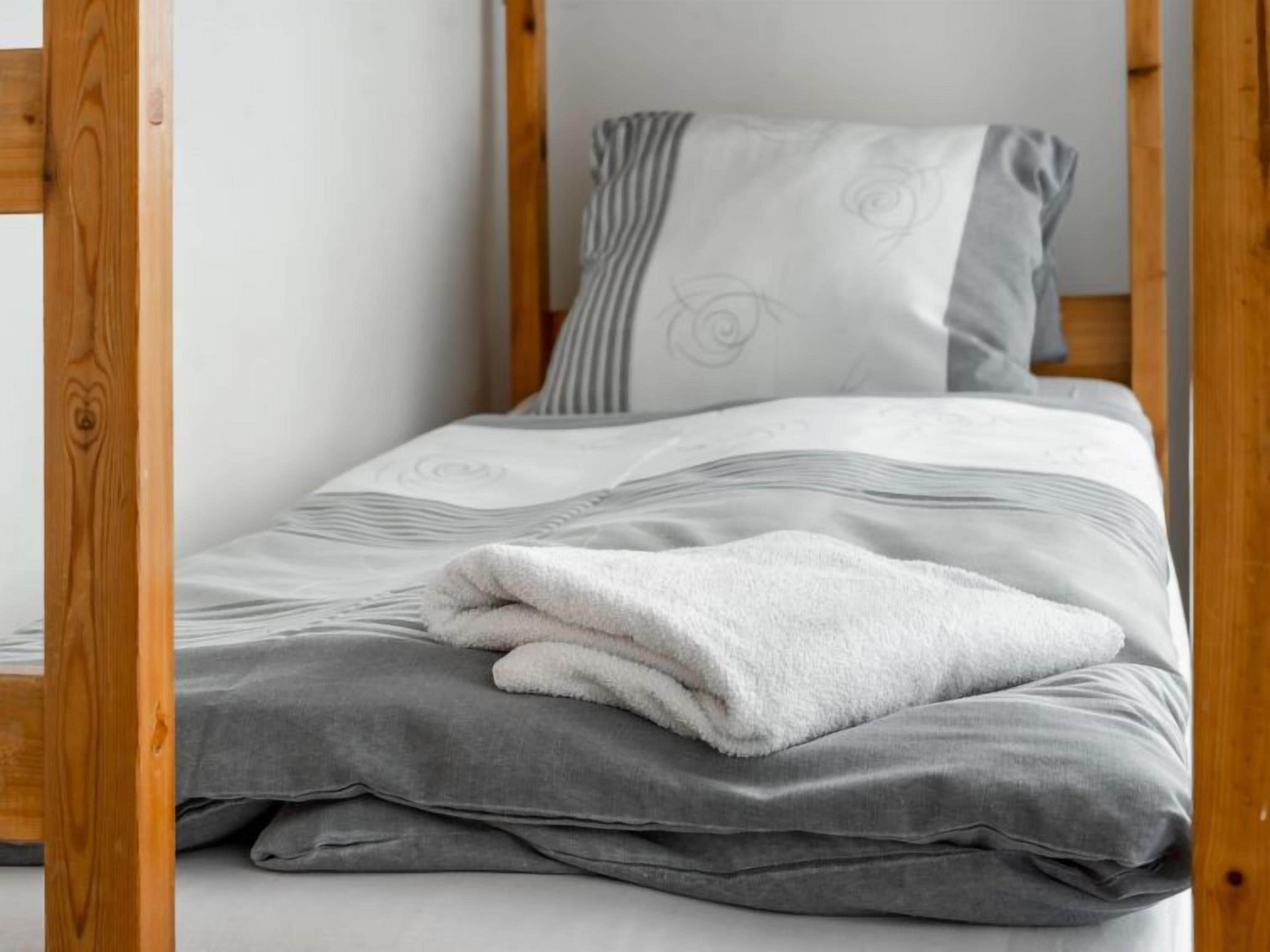 A single bed with a grey and white patterned bedspread, a white fluffy pillow, and a grey blanket.