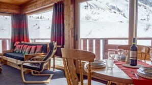 Una habitación acogedora con techo de madera, un sofá con cojines de colores, una mesa de comedor preparada para dos personas y vistas a un paisaje montañoso nevado.