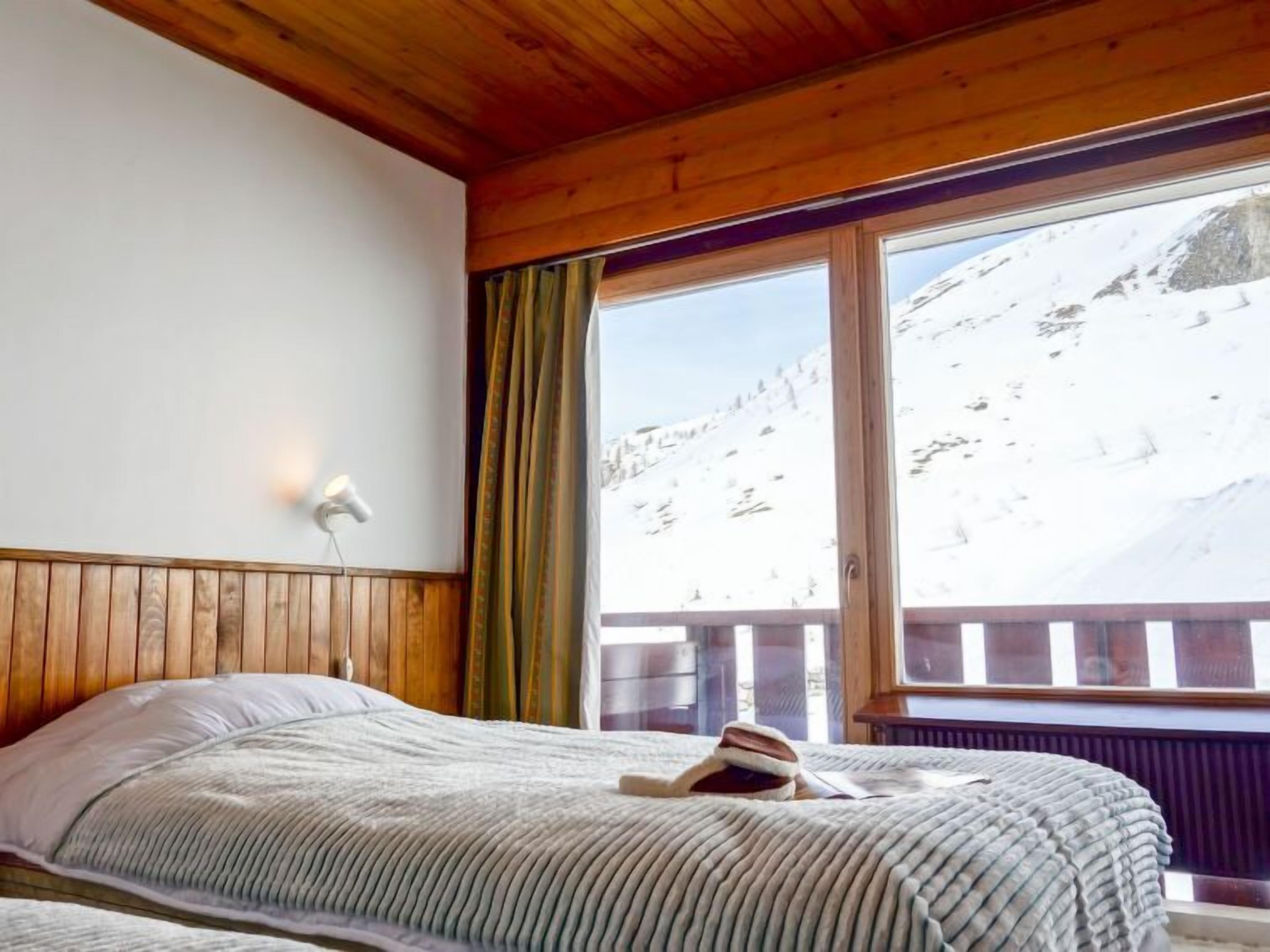 Une chambre avec un plafond en bois, un lit recouvert d’une couverture à rayures, une fenêtre donnant sur une montagne enneigée et une table de chevet avec une lampe.