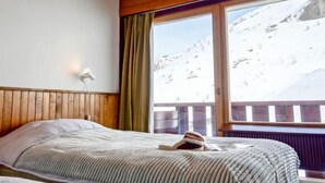 Un dormitorio con techo de madera, una cama con manta a rayas, una ventana con vista a una montaña nevada y una mesita de noche con lámpara.