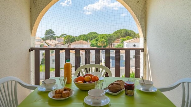 Un balcón con una mesa preparada para el desayuno, que incluye croissants, fruta y una botella de jugo, con vista a una zona residencial.