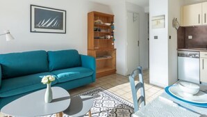 Ein modernes Wohnzimmer mit einem petrolfarbenen Sofa, einem kleinen runden Tisch mit Blumen und einem Holzregal, sowie einer Küchenzeile mit Schränken und einem Geschirrspüler.