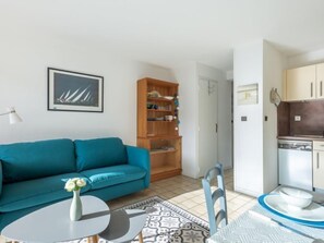 A modern living room with a teal sofa, a small round table with a vase of flowers, a wooden bookshelf, and a kitchen area with cabinets and a dishwasher.