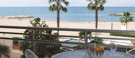 A balcony with a table set for two, overlooking a beach with palm trees and the sea.