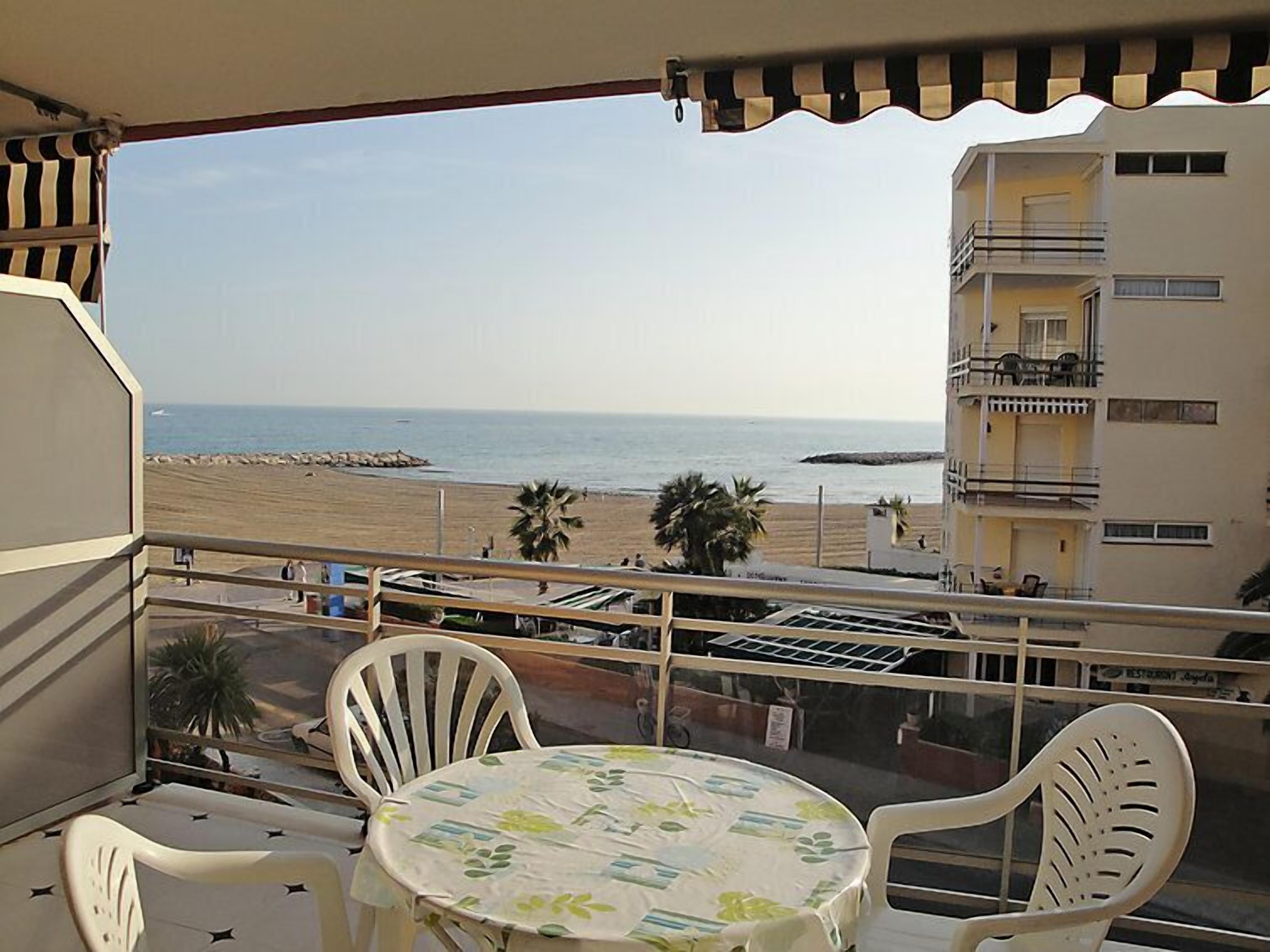 A balcony with a table and chairs, overlooking a beach and a building with balconies.