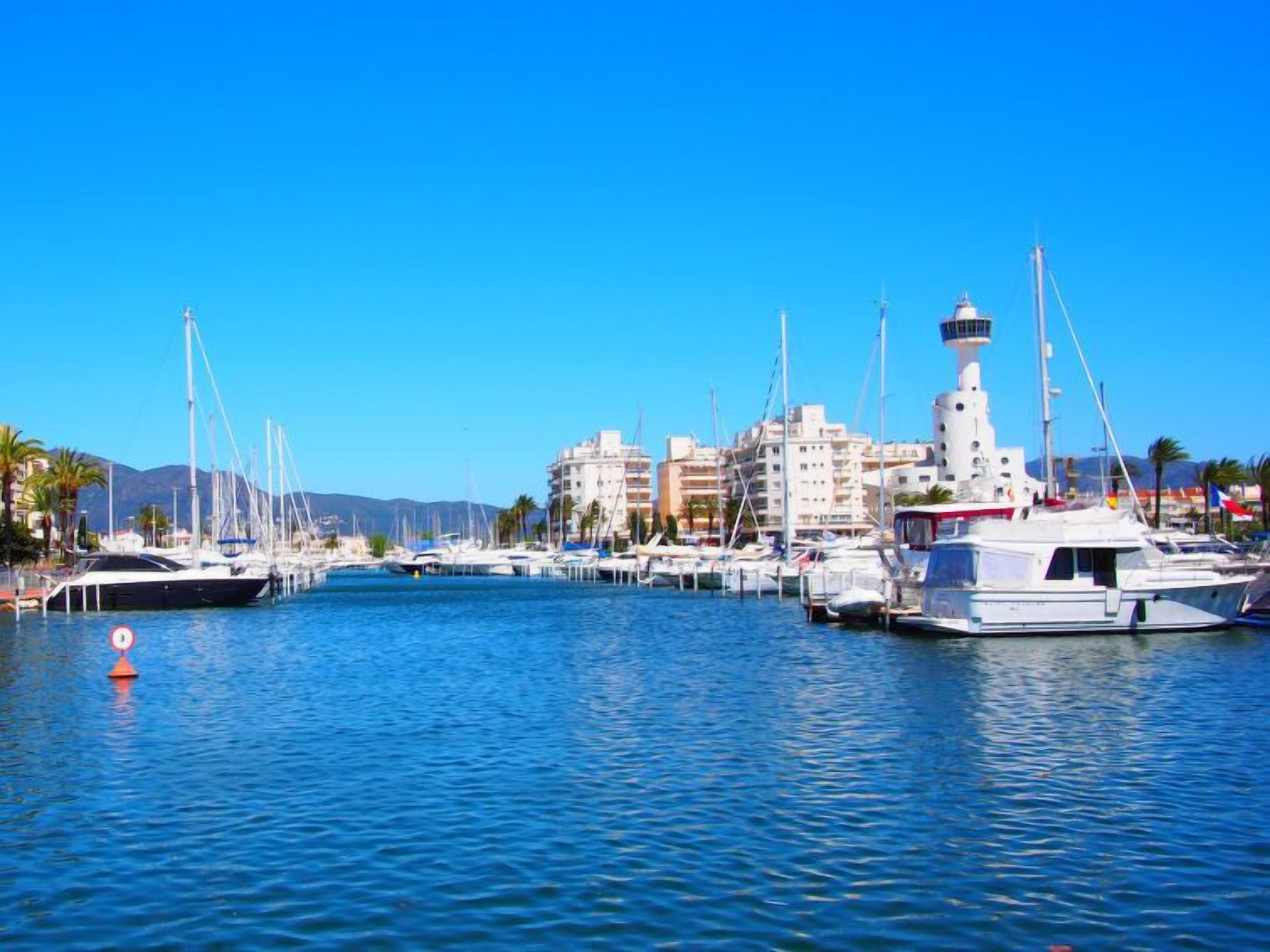 A marina with multiple yachts docked, a lighthouse, and buildings in the background.