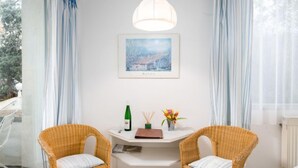 Una habitación con dos sillones de mimbre, una mesa blanca, un cuadro enmarcado, una botella de vino y un jarrón con flores.