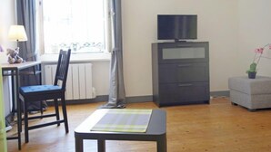 Huoneessa on puulattia, työpöytä, jossa on lamppu, tuoli, televisiotaulutaso ja pieni pöytä, jonka päällä on alusta.