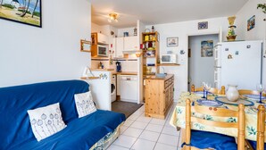 Ein kompakter Wohnbereich mit einem blauen Sofa, einem Esstisch mit Stühlen, einer Küchenzeile mit Mikrowelle und Backofen sowie einem kleinen Kühlschrank.