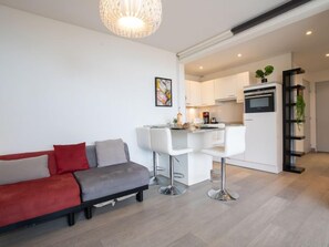 A modern living room with a sofa, a dining area with bar stools, a kitchen with white cabinets, and a built-in oven.