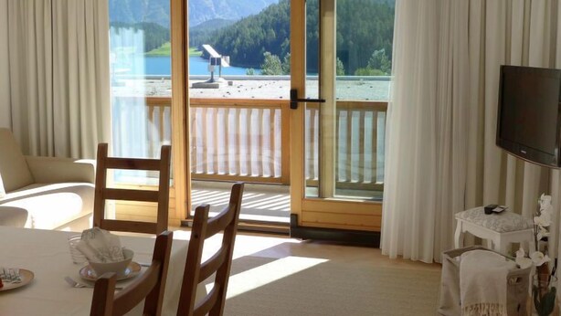 Ein Hotelzimmer mit einer großen Schiebetür zu einem Balkon mit Bergblick. Es gibt ein Bett, einen Sessel, einen kleinen Tisch und einen Fernseher.