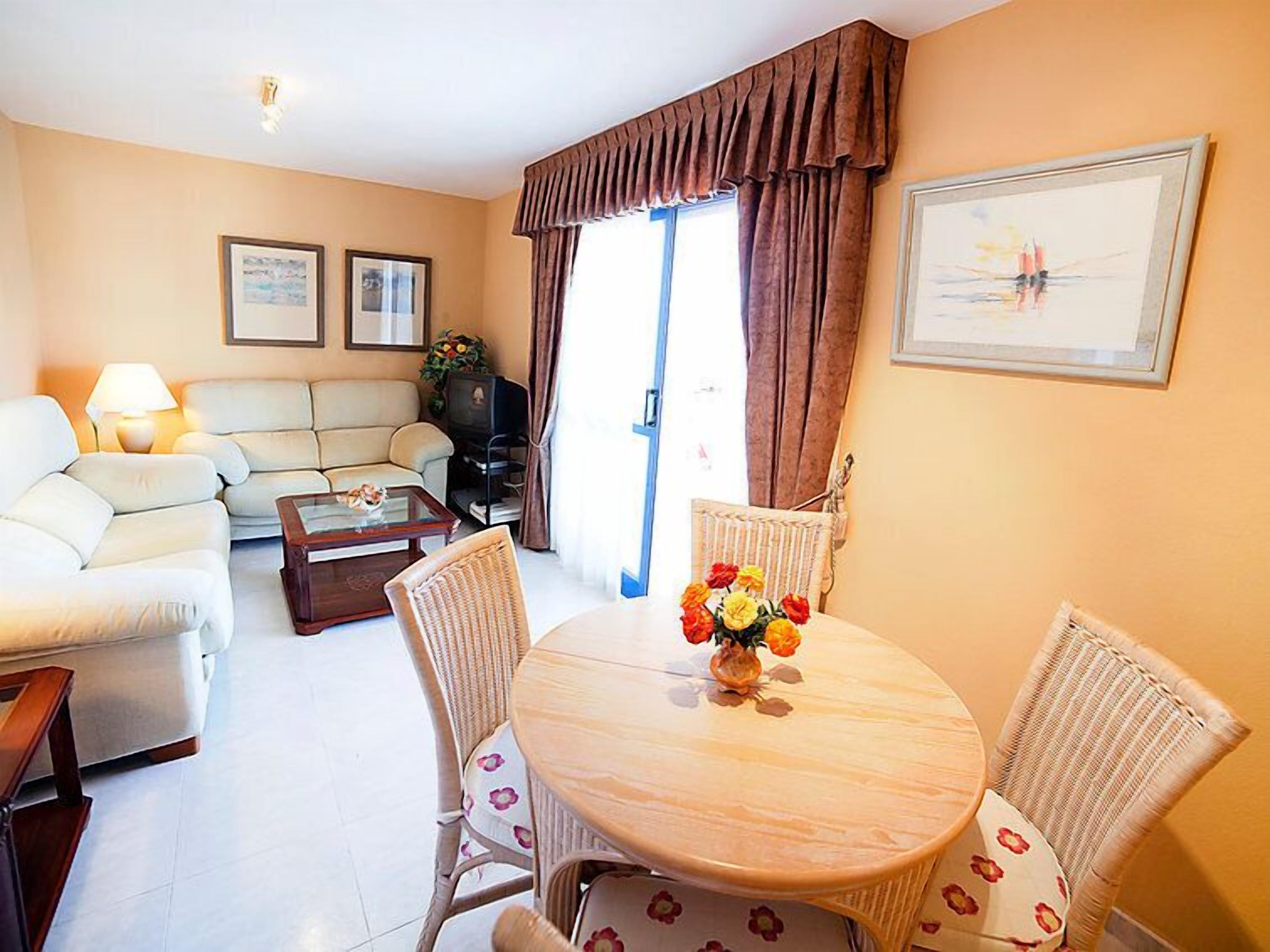 Un salon comprenant un canapé, une table basse, une table à manger avec des chaises, un téléviseur et une fenêtre avec des rideaux.