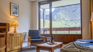 Ein gemütliches Zimmer mit einem Holzstuhl, einem kleinen Tisch mit einer Flasche und Gläsern, einem Fernseher und einem großen Fenster mit Blick auf die Berge.