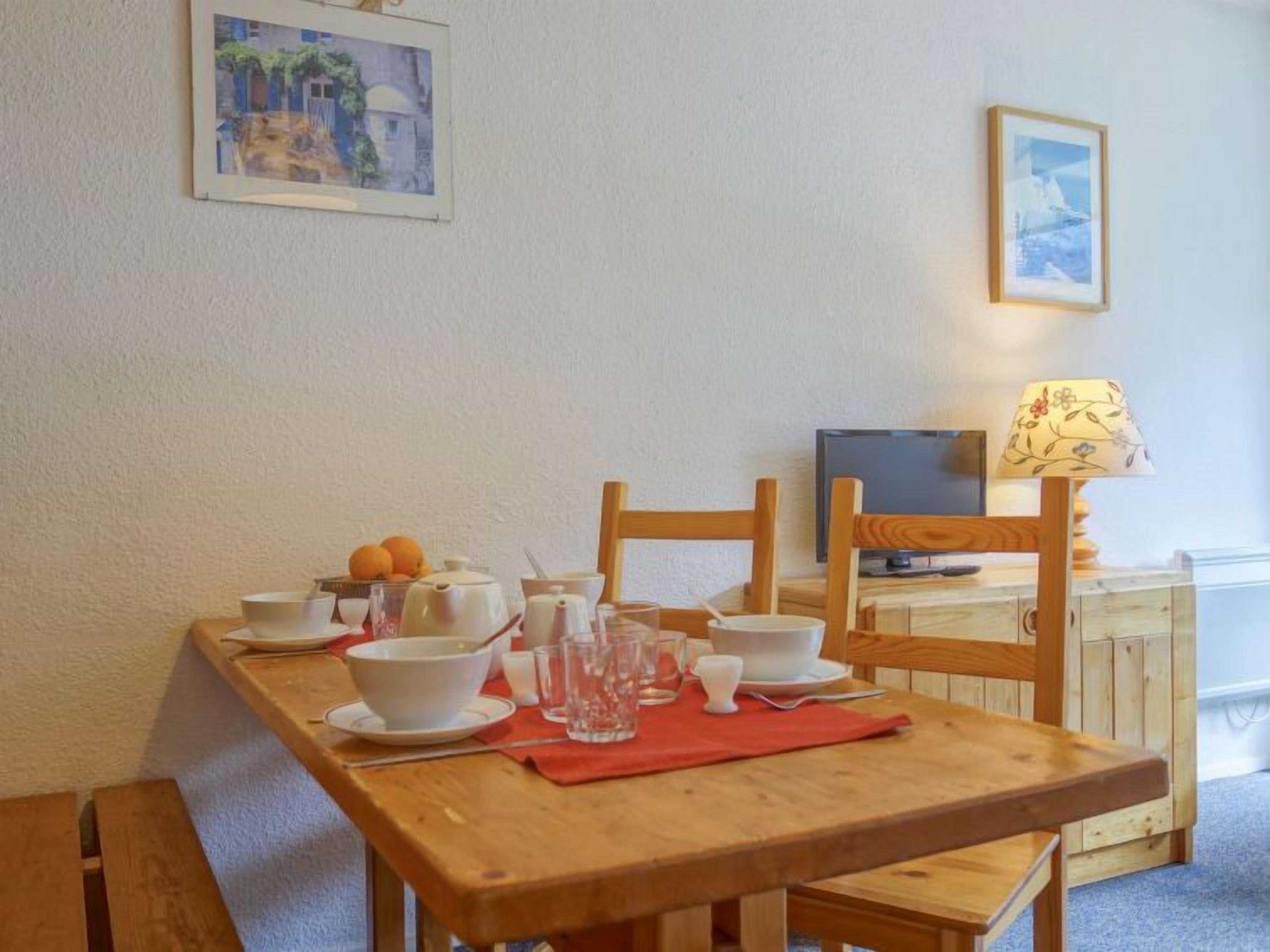 Een eetgedeelte met een houten tafel gedekt voor twee personen, een televisie en een lamp.