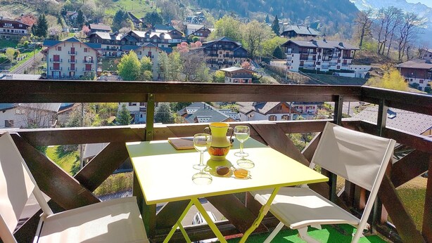 Un balcone con un tavolo apparecchiato per due, con vista su un borgo con case e montagne sullo sfondo.