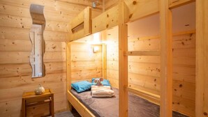 Quarto de cabana de madeira com beliches, uma mesinha e uma janela.